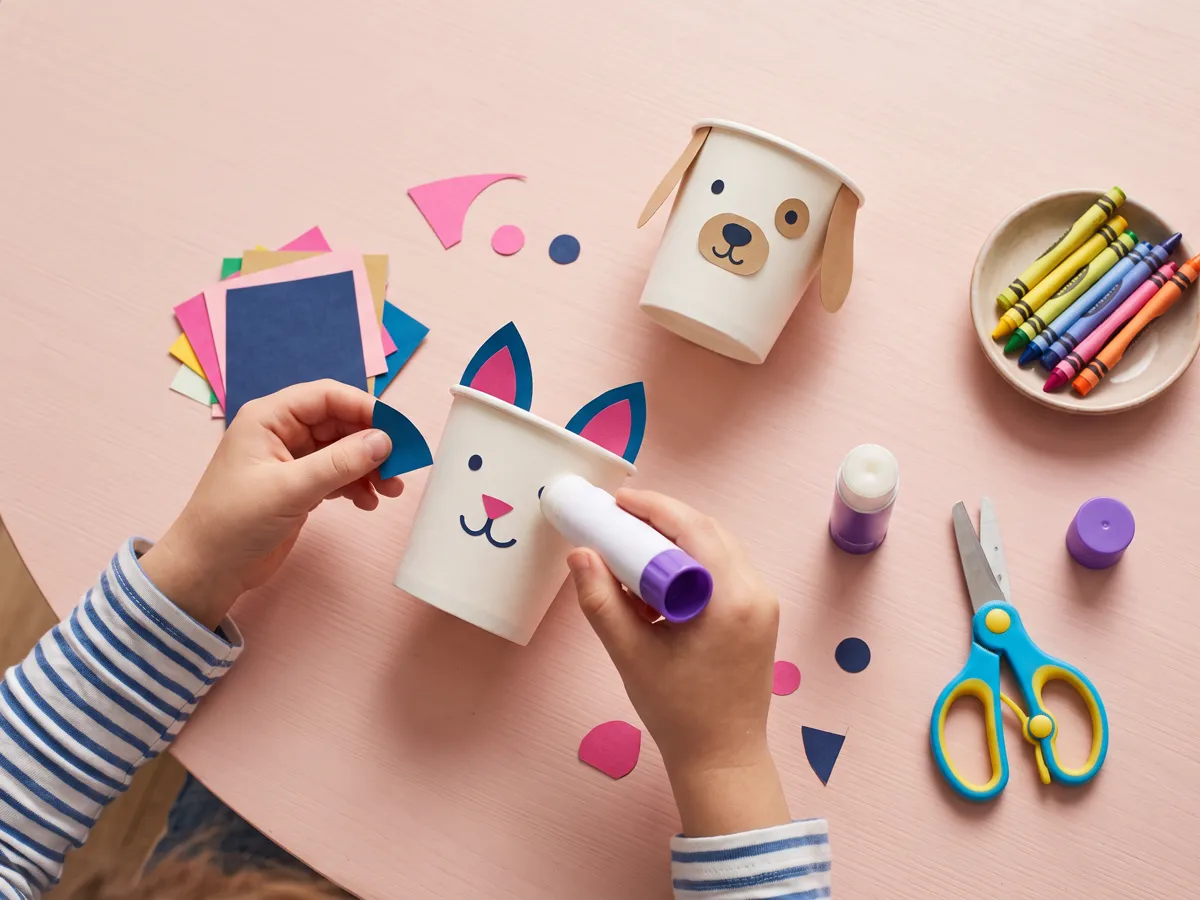 Paper-cup animal craft in progress with glued paper ears, crayons and safety scissors